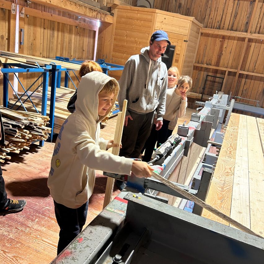 Ein junger Junge in einer Kapuzenjacke bedient eine große Holzbearbeitungsmaschine, wobei ein Mann und zwei Mädchen hinter ihm beobachten. Sie befinden sich in einem Raum mit Holzwänden und einem Boden, auf dem Holzbretter auf blauen Metallregalen gestapelt sind.