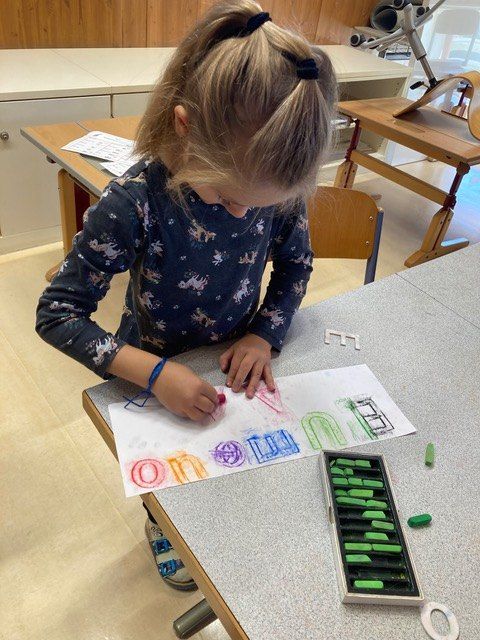 Ein kleines Mädchen malt in einem Klassenzimmer, konzentriert auf ein Stück Papier mit Buchstaben und Zahlen. Sie hat eine Schachtel mit farbigem Kreidestift auf dem Tisch.