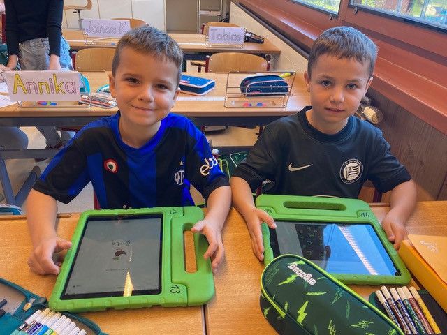 Zwei junge Jungen sitzen an einem Schreibtisch mit Tablets vor ihnen. Sie tragen Sporttrikots und halten jeweils ein Tablet in der Hand.