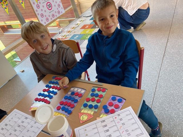 Zwei Jungen sitzen in einem Klassenzimmer an einem Tisch, lächeln und beschäftigen sich mit einem Spiel. Der Tisch hat pädagogische Materialien und nummerierte Karten. Ein Junge trägt einen blauen Pullover.