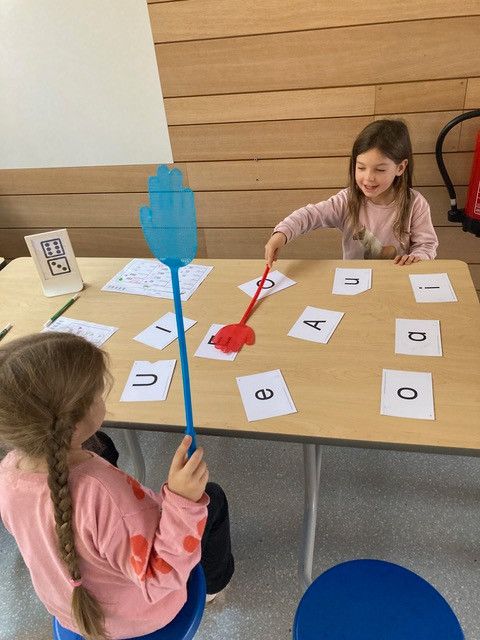 Zwei Mädchen sitzen an einem Tisch mit Buchstaben und Zahlen und benutzen eine Netz und einen Stock, wahrscheinlich spielen sie ein pädagogisches Spiel. Der Tisch ist mit Buchstaben und Zahlen bedeckt. In der Ecke steht ein Feuerlöscher.