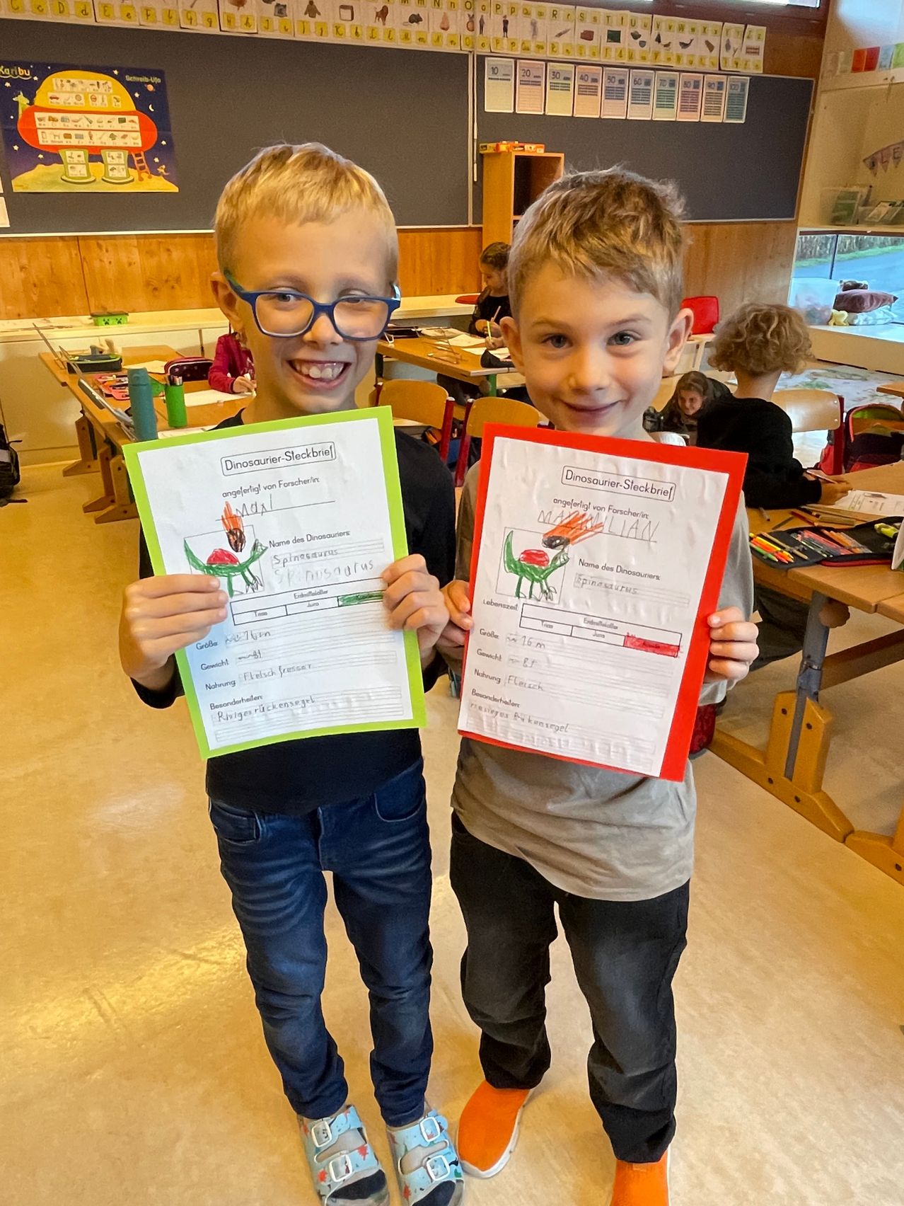 Zwei Jungen mit Brille posieren mit ihren Dinosaurier-Zeichnungen in einem Klassenzimmer. Sie halten bunte Papiere mit Dinosaurier-Zeichnungen.