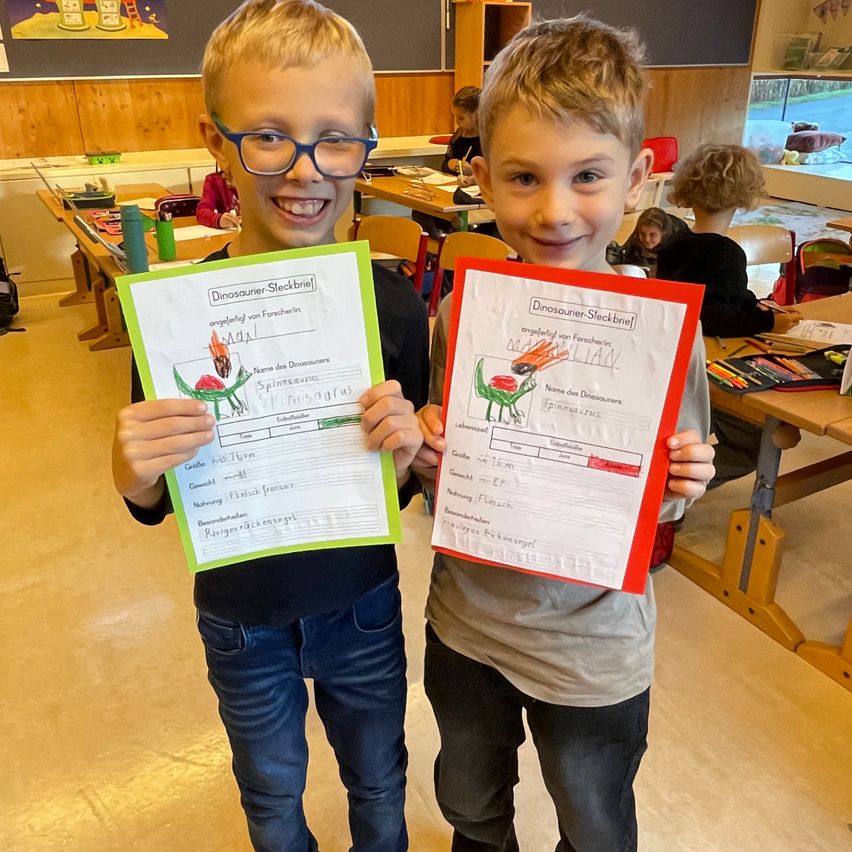 Zwei Jungen mit Brille posieren mit ihren Dinosaurier-Zeichnungen in einem Klassenzimmer. Sie halten bunte Papiere mit Dinosaurier-Zeichnungen.