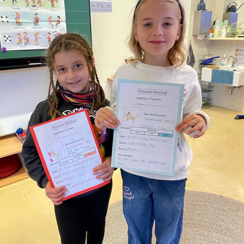 Zwei Mädchen in einem Klassenzimmer, eines hält ein Zertifikat mit einem Dinosaurier-Zeichnung und das andere ein Zertifikat mit einem bunten Design. Sie lächeln beide und schauen in die Kamera. Hinter ihnen befindet sich eine Tafel mit einigen Zeichnungen und Texten. Auf der rechten Seite gibt es ein Waschbecken mit einem Wasserhahn und einem Regal mit einigen Gegenständen.