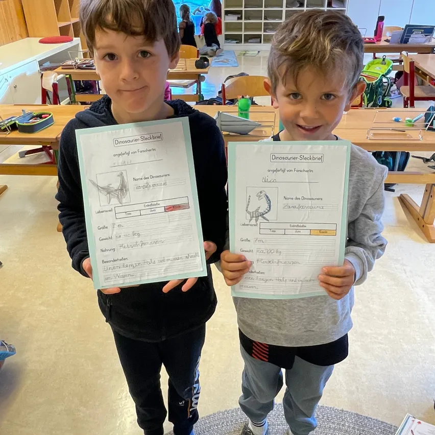 Zwei Jungen im Klassenzimmer halten Urkunden mit Dinosaurierzeichnungen und stehen auf einem Teppich. Einer hält eine Urkunde mit dem Namen 'Dinosaurier-Skizzenbrief'.