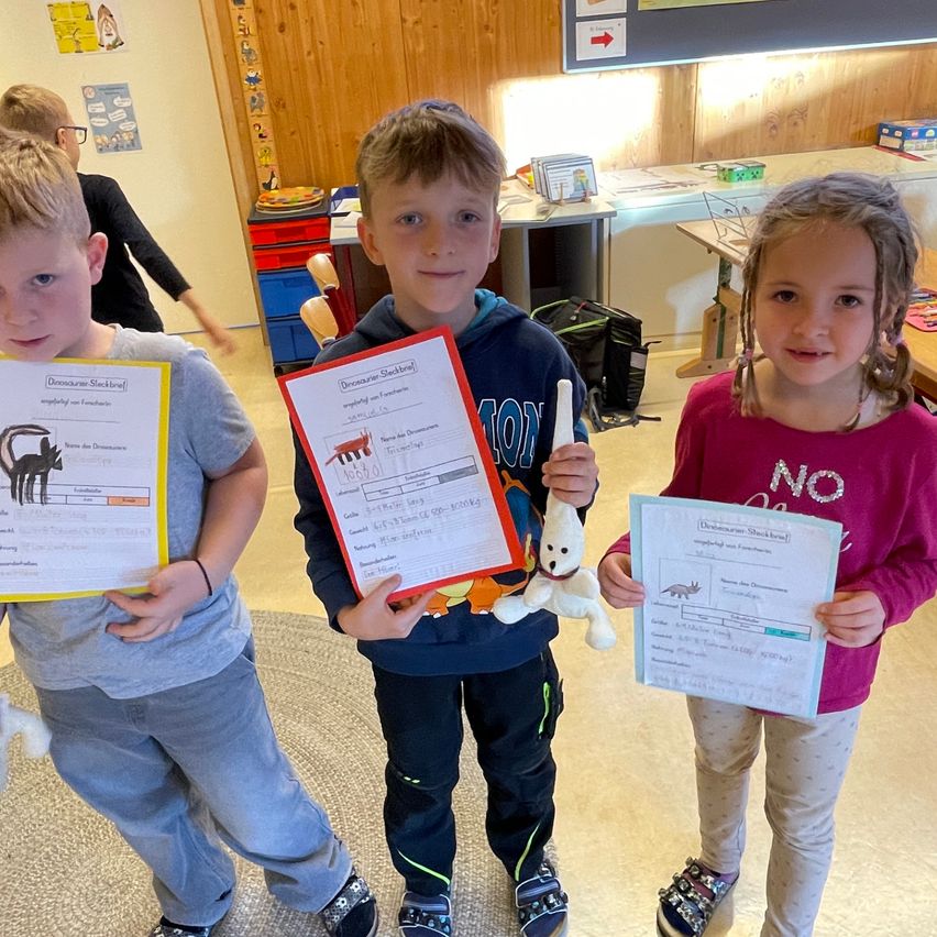 Drei Kinder halten Urkunden in der Hand und stehen in einem Klassenzimmer. Sie lächeln und posieren für ein Foto, wobei ein Kind einen Plüschtier hält.