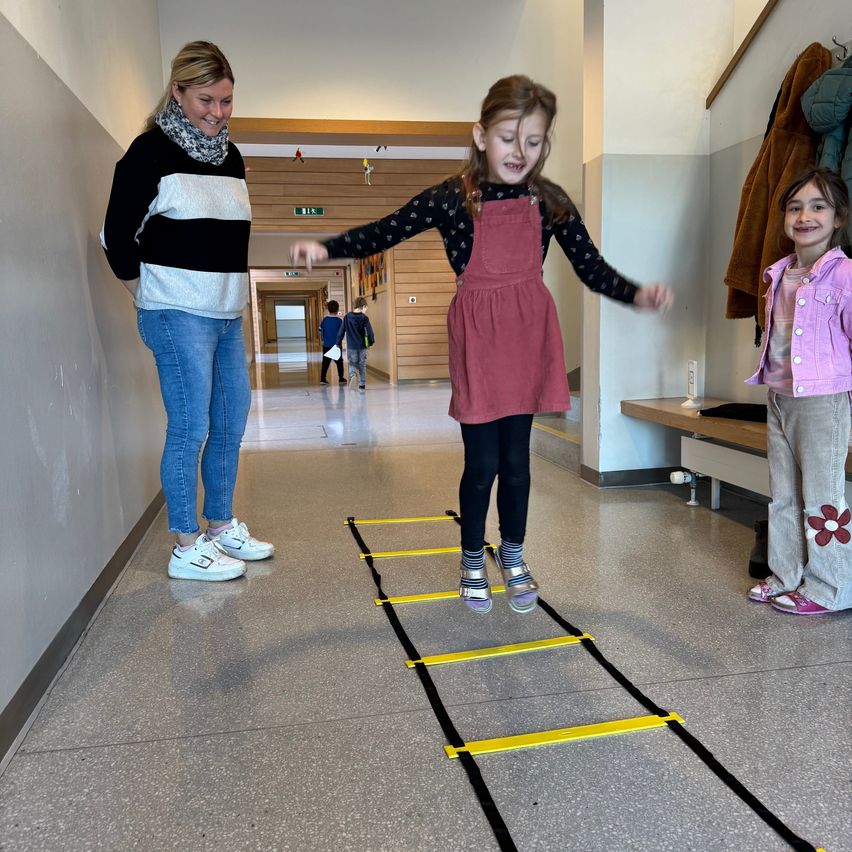 Eine Frau und zwei junge Mädchen springen auf einer gelben Leiter in einem Flur. Ein anderes Kind geht im Hintergrund.