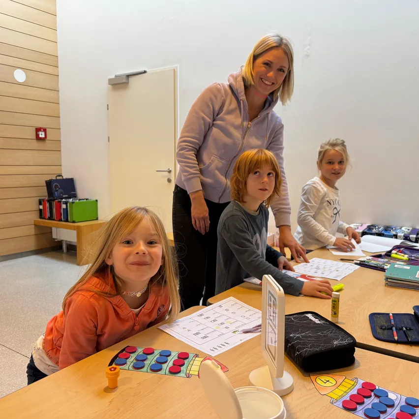 Eine lächelnde Frau und drei Kinder sitzen an einem Tisch in einem Klassenzimmer. Die Kinder haben verschiedene Lernmaterialien auf dem Tisch, einschließlich eines Spielbretts.