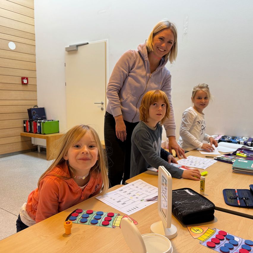 Eine lächelnde Frau und drei Kinder sitzen an einem Tisch in einem Klassenzimmer. Die Kinder haben verschiedene Lernmaterialien auf dem Tisch, einschließlich eines Spielbretts.