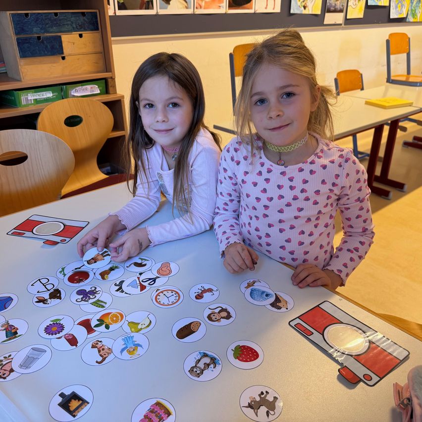 Zwei junge Mädchen sitzen an einem Tisch und lächeln, während sie mit Lernkarten spielen. Auf den Karten sind verschiedene Objekte und Buchstaben abgebildet. Sie befinden sich in einem Klassenzimmer mit Holzstühlen und -tischen.