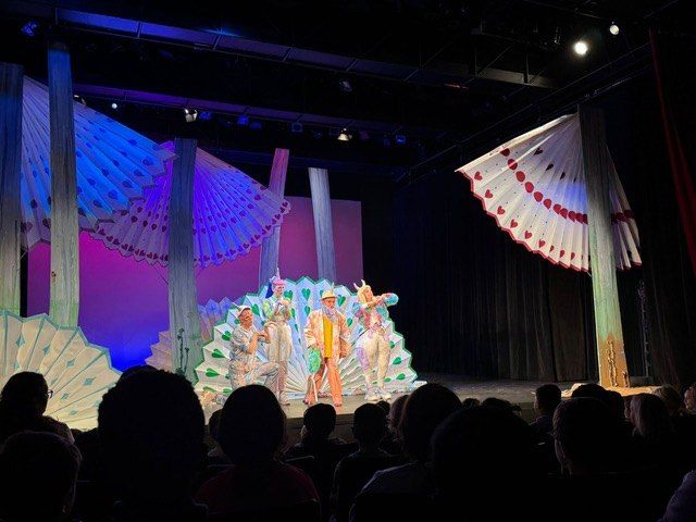 Eine farbenfrohe Bühnenaufführung mit Pfauen-Thema. Vier Darsteller stehen unter leuchtenden Lichtern und einem dekorativen Hintergrund auf der Bühne. Ein Publikum beobachtet von vorne.