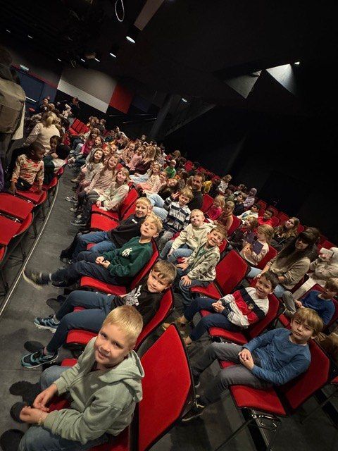 Ein Theater voller Kinder und Erwachsener, die auf roten Stühlen sitzen. Einige lächeln und andere schauen etwas an.
