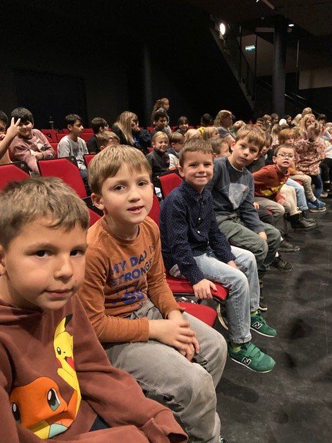 Eine Gruppe von Kindern sitzt in roten Stühlen in einem Theater und schaut mit aufmerksamen Gesichtern einer Aufführung zu.