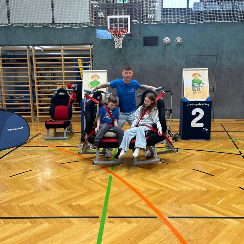 Ein Mann mit Tätowierungen steht mit zwei Kindern in Trainingssitzen in einer Turnhalle, ein Basketballkorb im Hintergrund.