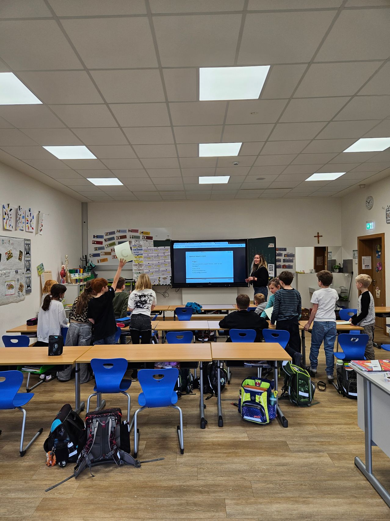 Ein Klassenzimmer mit Schülern, die um Tische herum sitzen, eine Lehrerin steht vor einer großen Leinwand, und einige Schüler stehen und schauen auf den Bildschirm. Das Klassenzimmer hat einen Holzboden und weiße Wände.