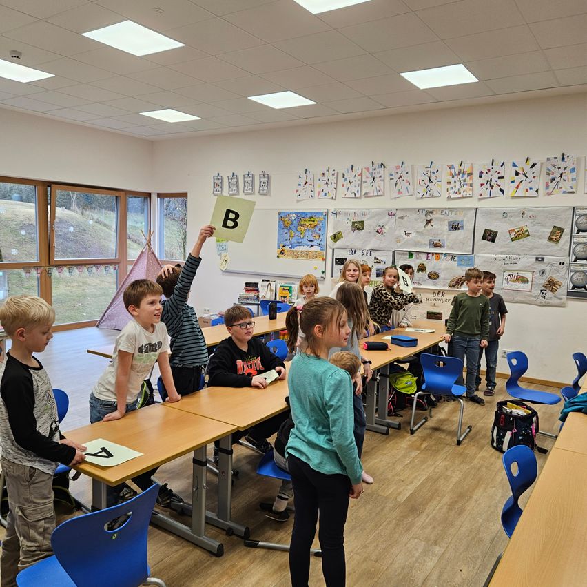 Ein Klassenzimmer mit Kindern, die stehen und Buchstabenkarten halten, einschließlich eines Jungen, der den Buchstaben B hält. Andere Kinder sitzen an Schreibtischen. Der Raum hat Bulletin Boards, Tische und Stühle.