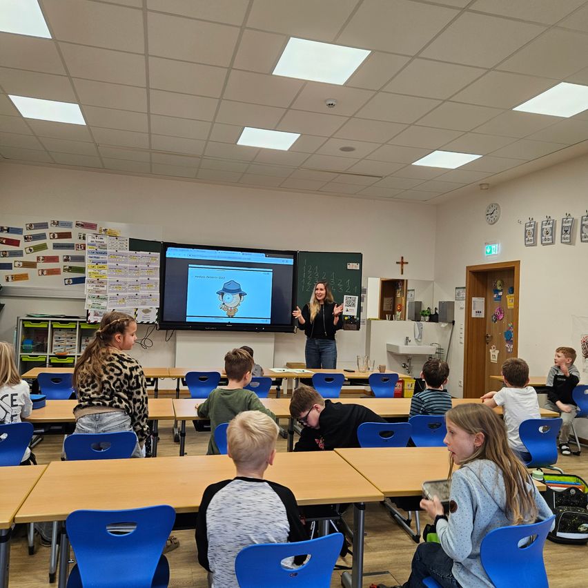 Eine Lehrerin steht vor einer Klassenpräsentation auf einem Bildschirm und erklärt eine Cartoonfigur aufmerksamen Schülern, die an Tischen in einem Klassenzimmer sitzen.