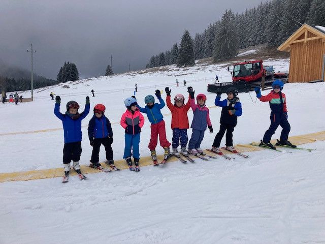 Eine Gruppe von Kindern in Skiausrüstung posiert für ein Foto auf einer verschneiten Piste mit einem Schneepflug im Hintergrund.