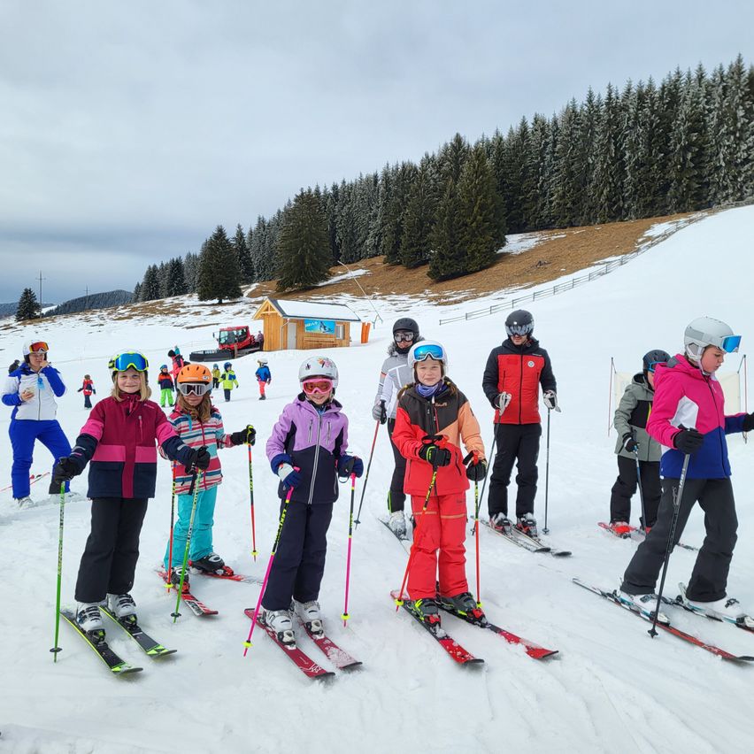 Eine Gruppe von Kindern und Erwachsenen skifahrer auf einem verschneiten Hang, in Winterkleidung und Skiern. Im Hintergrund befindet sich ein kleines Gebäude und ein Maschinenfahrzeug.