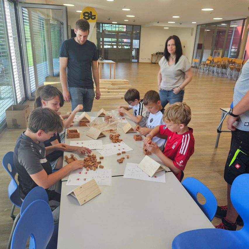 Mehrere Kinder sitzen um einen Tisch herum und bauen etwas. Ein Mann und eine Frau stehen hinter ihnen. Der Raum hat Holzböden, Glaswände und einen Tisch voller Stühle.