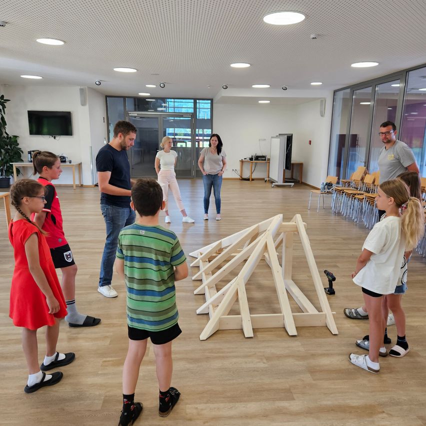 Eine Gruppe von Erwachsenen und Kindern versammelt sich um eine dreieckige Holzkonstruktion auf einem Holzboden. Sie nehmen wahrscheinlich an einem Workshop oder einer Klasse teil.