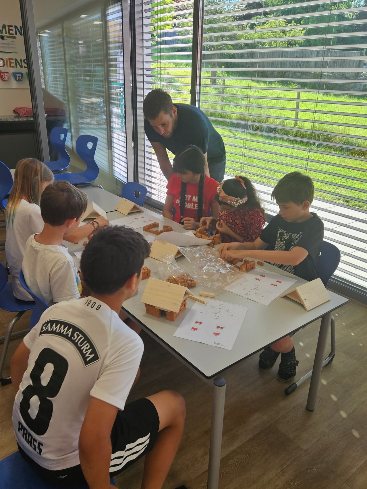 Eine Gruppe von Kindern arbeitet an einem Tisch unter einem Glasfenster mit Jalousien an Holzarbeiten. Ein Erwachsener steht hinter ihnen und leitet sie.