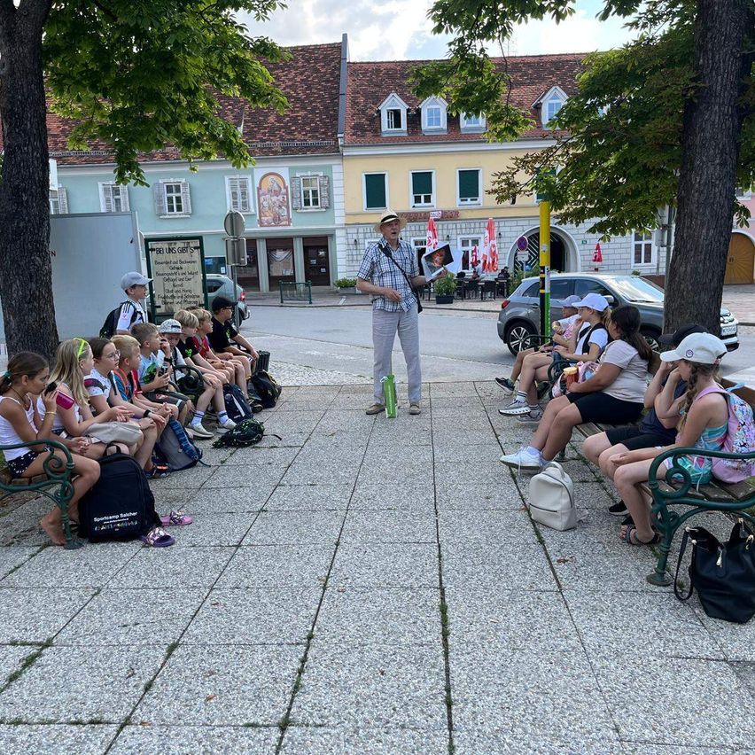 Ein Mann spricht mit einer Gruppe von Kindern, die auf Bänken auf einem Stadtplatz sitzen. Im Hintergrund befinden sich Bäume, Gebäude und geparkte Autos.