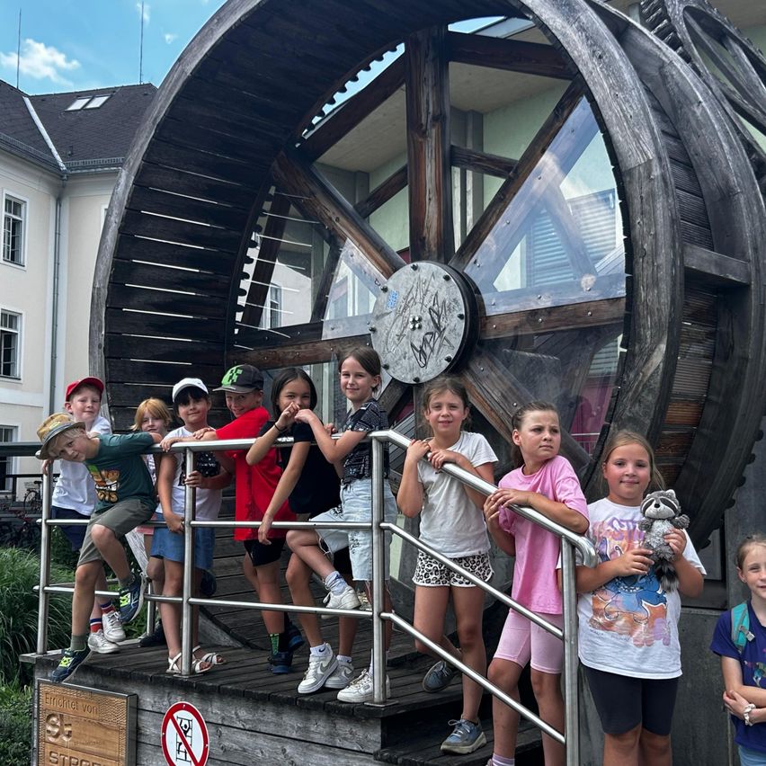 Eine Gruppe von Kindern posiert vor einem großen hölzernen Rad mit Graffiti in einem Park. Sie lächeln und halten sich an den Geländern fest.