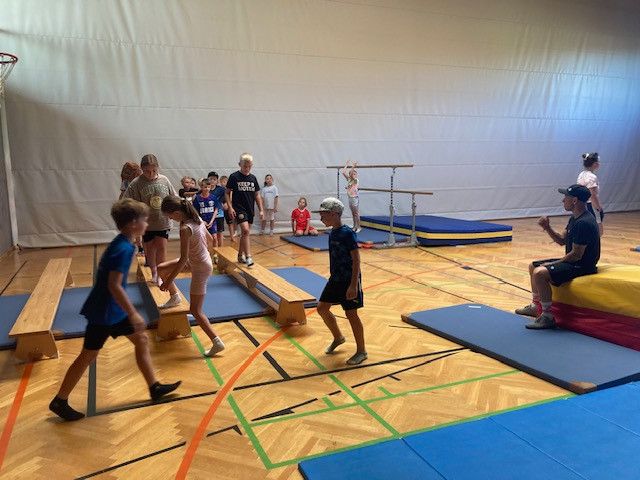 Eine Gruppe von Kindern und Erwachsenen in einer Turnhalle, die verschiedene Übungen auf Matten und Holzbalken übt.