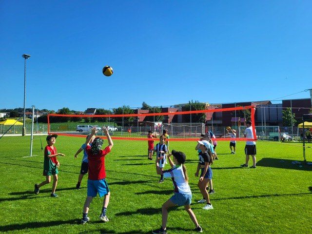 Eine Gruppe von Kindern spielt Volleyball auf einem grünen Feld unter einem klaren blauen Himmel. Sie sind in zwei Teams aufgeteilt.