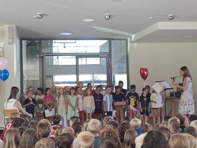 A group of children in a classroom are standing in line, some holding balloons and papers, while a woman with a guitar and a music stand stands in front of them.