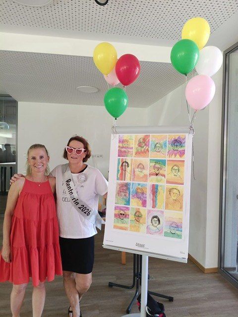 Two women stand next to a poster with colorful drawings of faces, one wearing a sash that says 'Rein in 2026'. Balloons are floating above the poster.