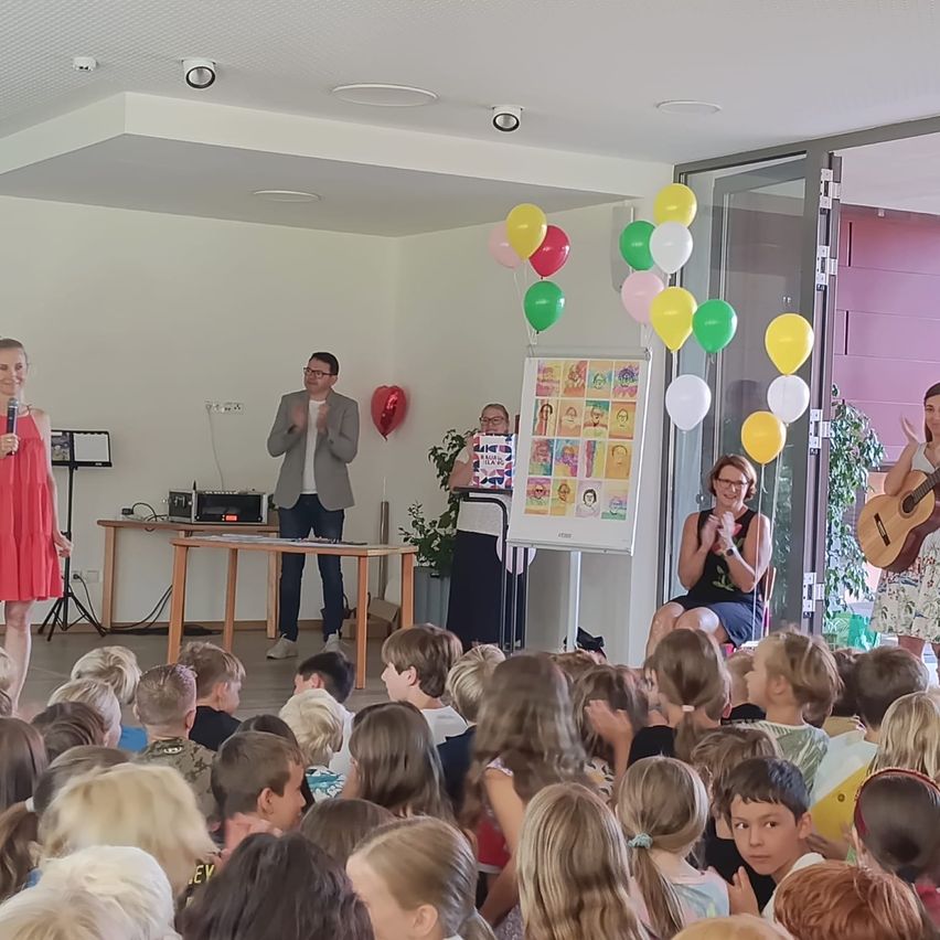 A group of people, including children, are gathered in a room for an event. A woman in a red dress stands on the left, and a man in a gray suit stands in the center. A whiteboard with pictures is behind them, and colorful balloons are floating in the air.