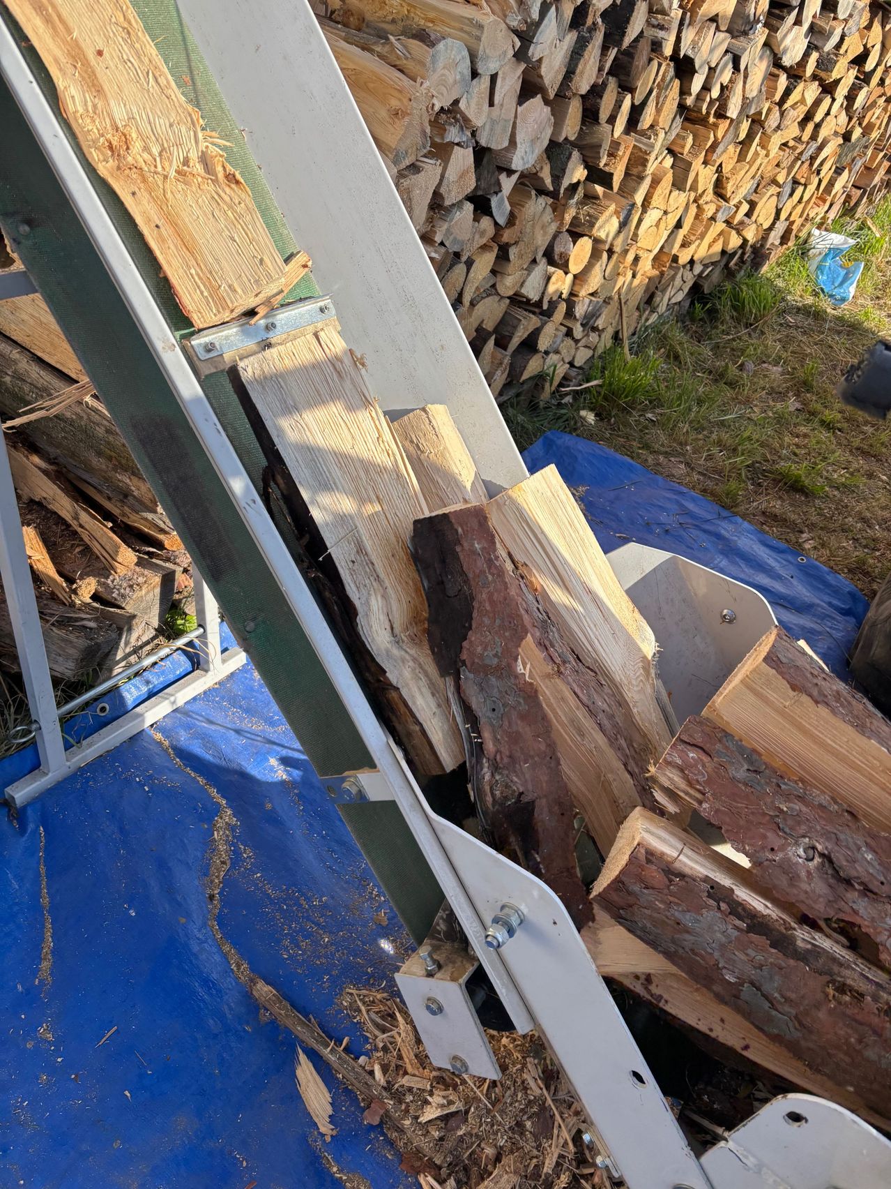 Ein Metallrahmen ist mit geschnittenen Baumstämmen beladen. Hinter ihm sind Holzstapel auf einer blauen Plane gestapelt, mit Gras und einer Plastiktüte in der Nähe.