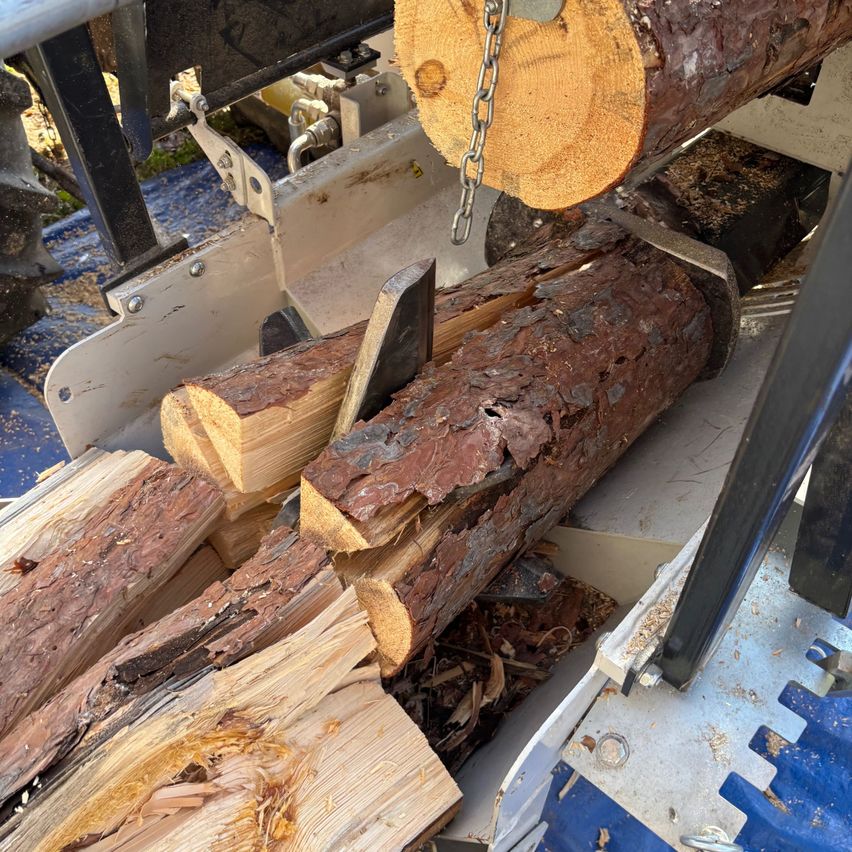 Ein Holzspalter schneidet einen Baumstamm in kleinere Stücke. Der Stamm wird von einer Metallkette gehalten, und die Sägeklinge ist sichtbar.