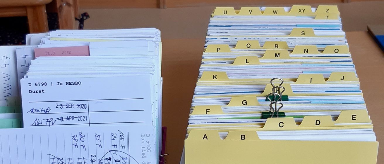 In einem Klassenzimmer stehen zwei Schachteln mit Karteikarten auf einem Schreibtisch. Eine Schachtel ist lila, die andere weiß. Die Schachteln sind mit Buchstaben beschriftet. Im Hintergrund befinden sich mit Büchern gefüllte Regale.