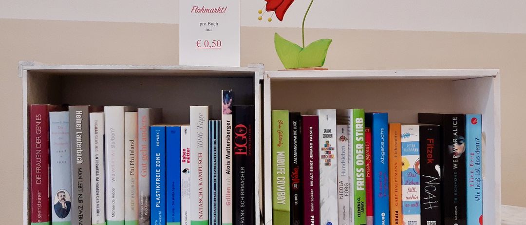 Ein Bücherregal mit mehreren Büchern und einem Schild, das 'Flohmarkt! pro Buch nur 0,50' sagt. Ein roter blumenförmiger Aufkleber ist an der Wand hinter dem Bücherregal.