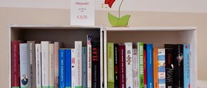 Ein Bücherregal mit mehreren Büchern und einem Schild, das 'Flohmarkt! pro Buch nur 0,50' sagt. Ein roter blumenförmiger Aufkleber ist an der Wand hinter dem Bücherregal.