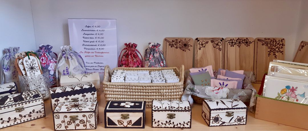 Eine Ausstellung dekorativer Holzkisten und Tüten, mit einem Schild, das Preise und Artikel auflistet. Körbe gefüllt mit kleinen Gegenständen und blumengeschmückte Holzbretter sind ebenfalls vorhanden.