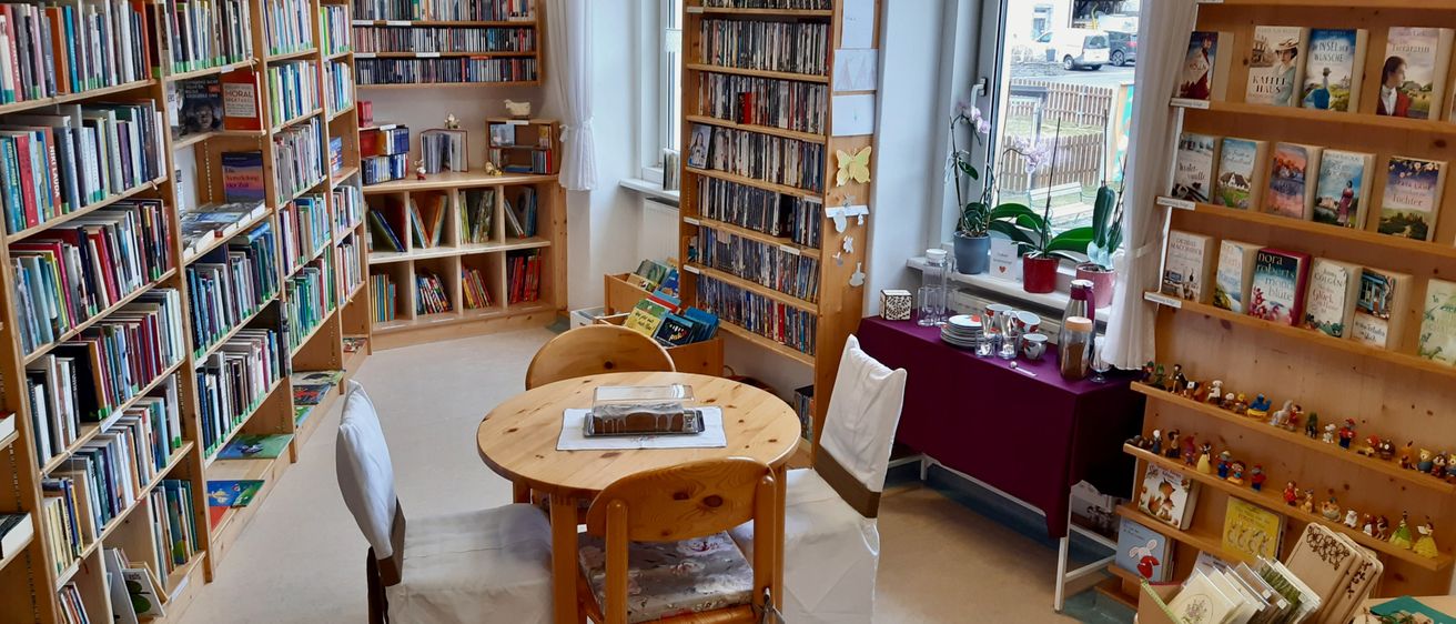 A room with a round wooden table with a cake covered in a glass container. A wooden chair is on the left. A wooden shelf is on the right with many books. A potted plant is on the window sill.