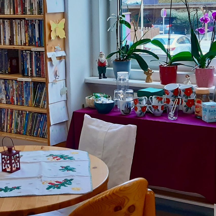 Ein gemütlicher Raum mit einem Holzregal voller DVDs, einem Tisch mit festlicher Tischdecke und einem Fenster mit Topfpflanzen und Blick auf ein Auto.