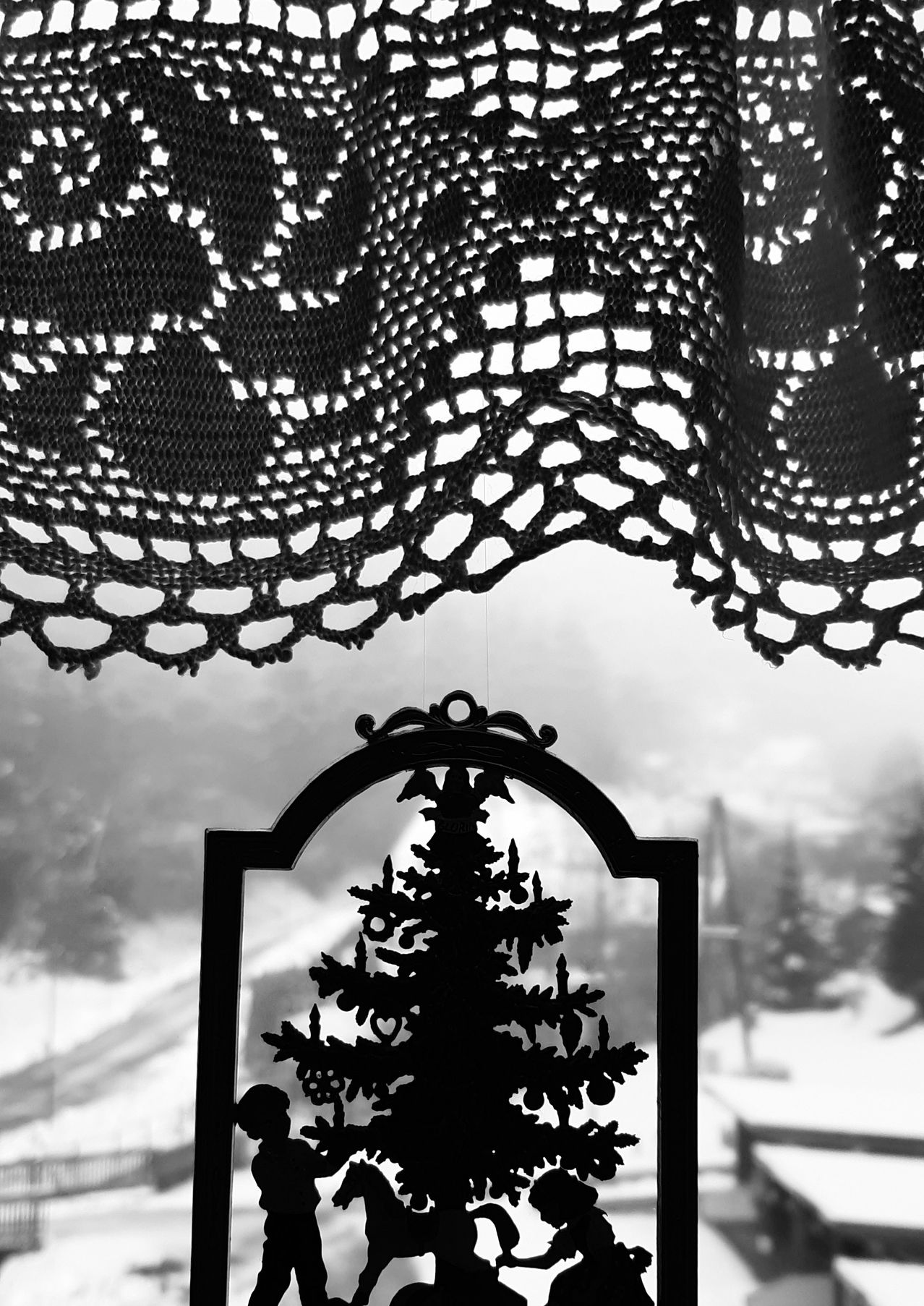 A black-and-white image features a lace curtain hanging over a window. Below it, a decorative silhouette of a Christmas tree is visible against a snowy backdrop.