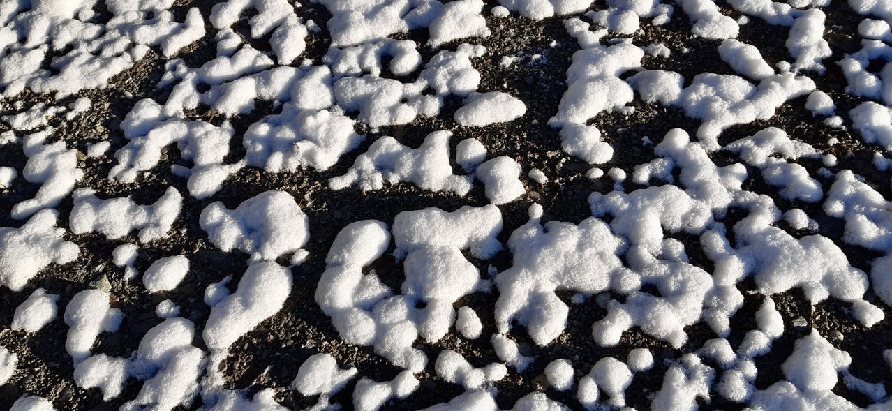 Eine Nahaufnahme eines verschneiten Bodens mit verschiedenen Formen und Größen von Schneekristallen. Der Schnee wird von Sonnenlicht beleuchtet, das Schatten auf den Boden wirft.