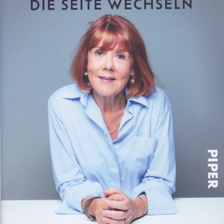 A woman with red hair is smiling and sitting on a white desk. She wears a light blue shirt and a necklace. The text 'DIE SEITE WECHSLEN' is above her.