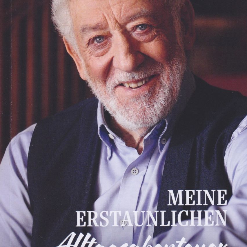 Ein lächelnder älterer Mann mit weißem Haar und Bart, der ein hellblaues Hemd und eine dunkle Weste trägt, posiert für die Kamera mit einem Buch mit dem Titel 'Meine Erstaunlichen Abenteuer'.