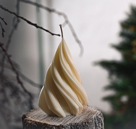 Eine weiße Kerze in Form eines Weihnachtsbaums steht auf einem Holzstamm mit kahlen Zweigen. Der Hintergrund ist unscharf, mit einem Weihnachtsbaum und roten Lichtern.