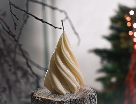 Eine weiße Kerze in Form eines Weihnachtsbaums steht auf einem Holzstamm mit kahlen Zweigen. Der Hintergrund ist unscharf, mit einem Weihnachtsbaum und roten Lichtern.