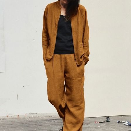 Eine Frau in einem schwarzen Oberteil und einer braunen Jacke geht auf einem Holzboden. Sie trägt braune Hosen, und die Szene zeigt verstreute Gegenstände auf dem Boden und eine weiße Wand im Hintergrund.