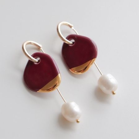 Ein Paar Ohrringe mit einem burgunderroten Mittelpunkt und goldener Einfassung, mit weißen Perlen. Die Ohrringe hängen an goldenen Haken.