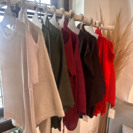 Eine Reihe von Pullovern in verschiedenen Farben, darunter weiß, beige, grün, rot und braun, hängt an einem Holzständer an einer weißen Wand.